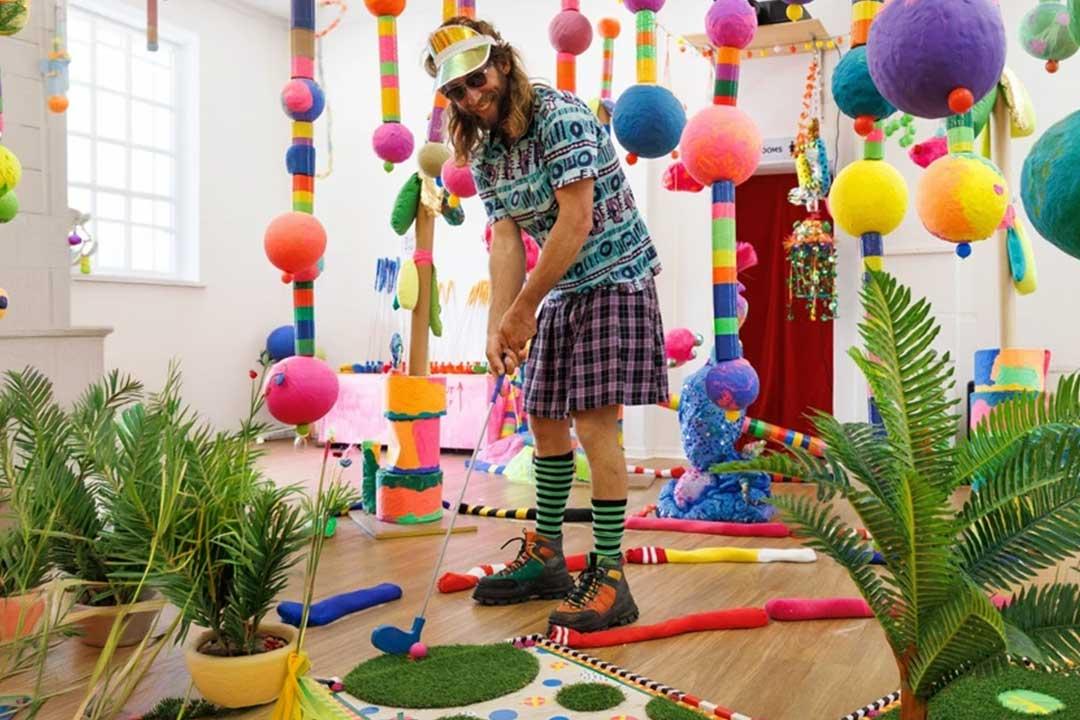 Bearded man in sunglasses plays whimsical indoor mini-golf amid colorful hanging balls, striped poles, tropical plants, and bright playful décor.