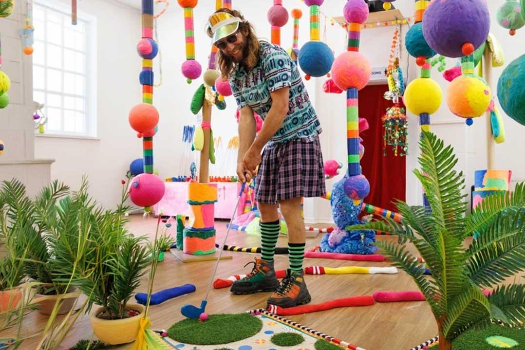 Bearded man in sunglasses and colorful clothes putts a golf ball in a whimsical indoor mini-golf room filled with pom-poms and plants.