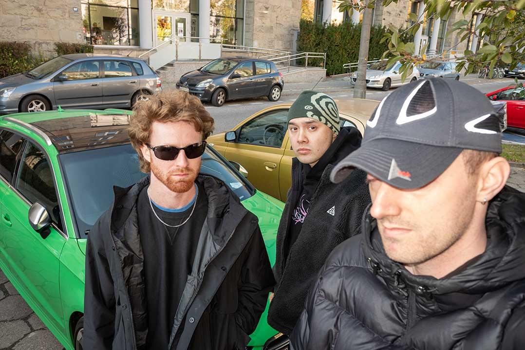 Three men pose beside a bright green car on a city street; sunglasses, beanies, and puffer jackets create a cool, urban mood.