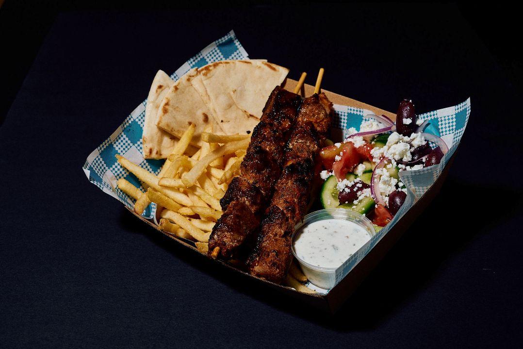 Tray of grilled meat skewers served with pita bread, hot chips and a Greek-style salad topped with feta cheese and olives.