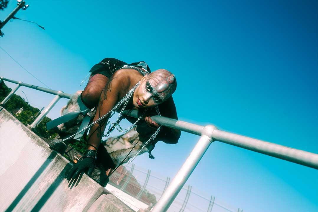 Tattooed punk person in leather and chains leans over a metal railing, staring intensely at the camera against a bright blue sky.