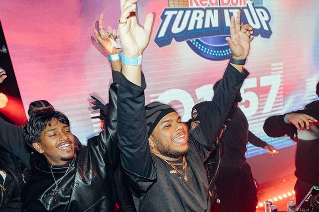 Smiling performers raise their hands onstage beneath a Red Bull Turn Up sign, surrounded by colorful lights and cheering people at a lively event.