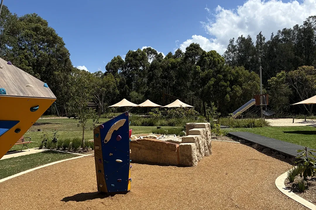 Outdoor playground with climbing structures, rocks, sandpit, and slide surrounded by grass, trees, and shade canopies under a partly cloudy sky.