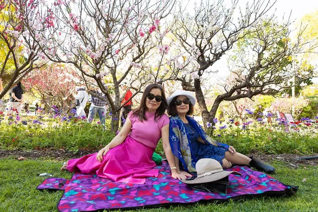 Two women in bright dresses relax on a colorful picnic blanket in a flower-filled park, smiling beneath blooming trees in a cheerful spring scene.