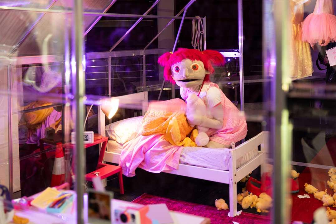 Pink-haired puppet doll sits on a small white bed in a colorful glass display, surrounded by plush toys and glowing purple lights.
