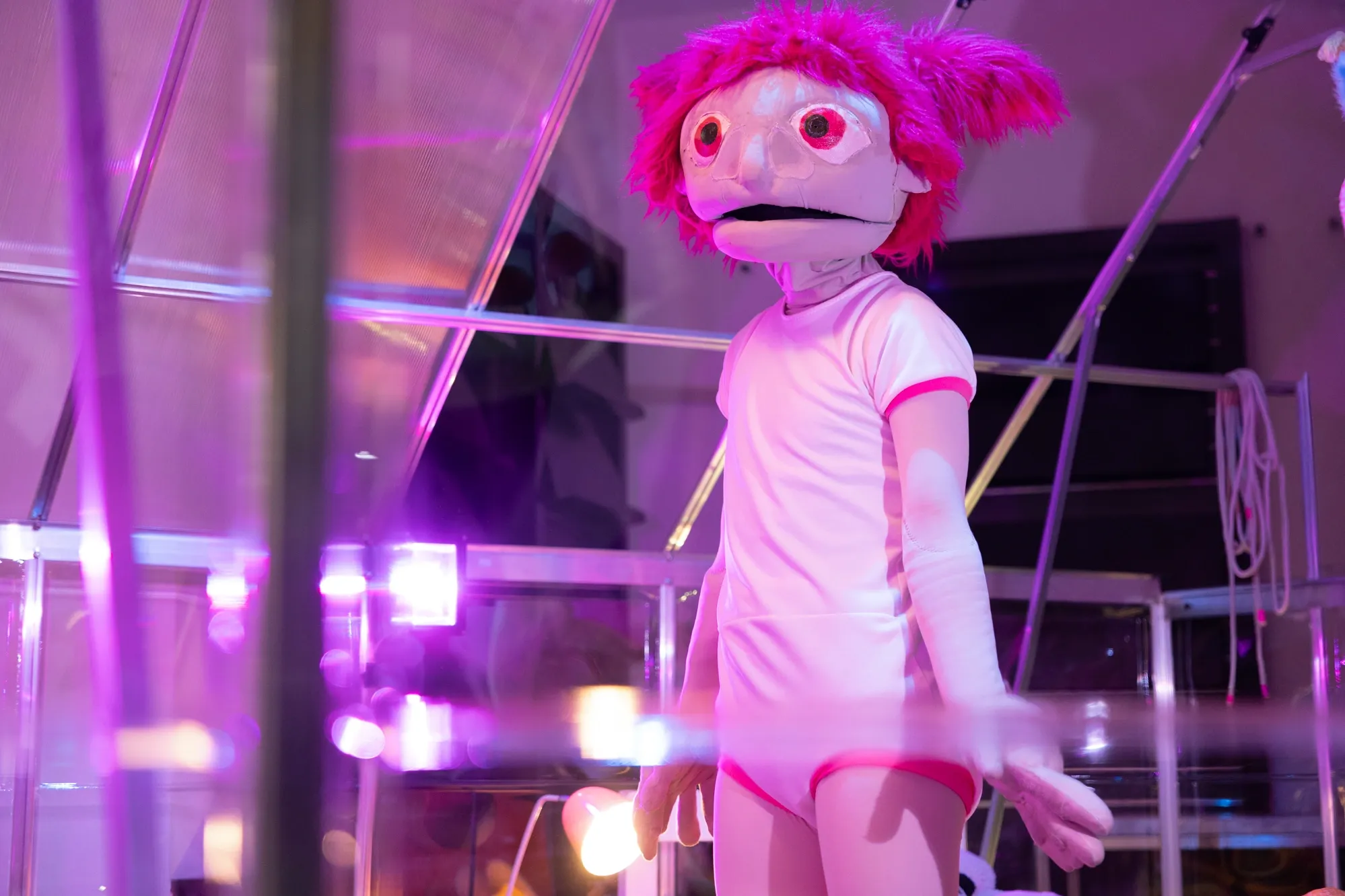 A large puppet with bright pink hair, oversized eyes and a white bodysuit stands inside a geometric structure illuminated by purple and pink lighting.