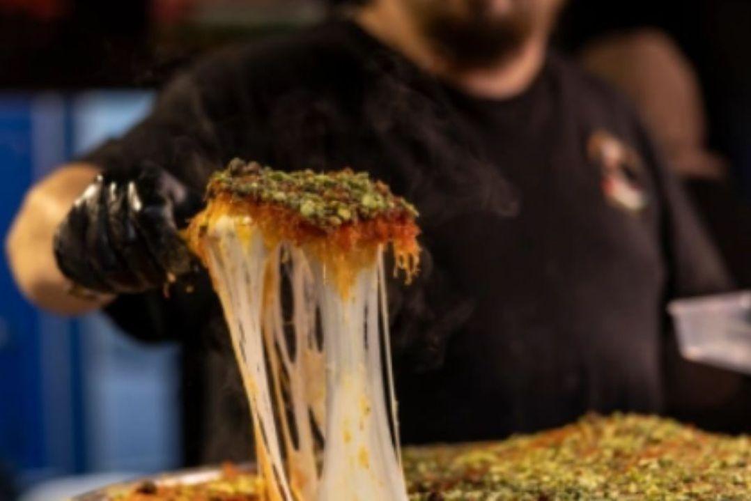 freshly made knafeh dessert being lifted from a tray, with long strands of melted cheese stretching from the slice.