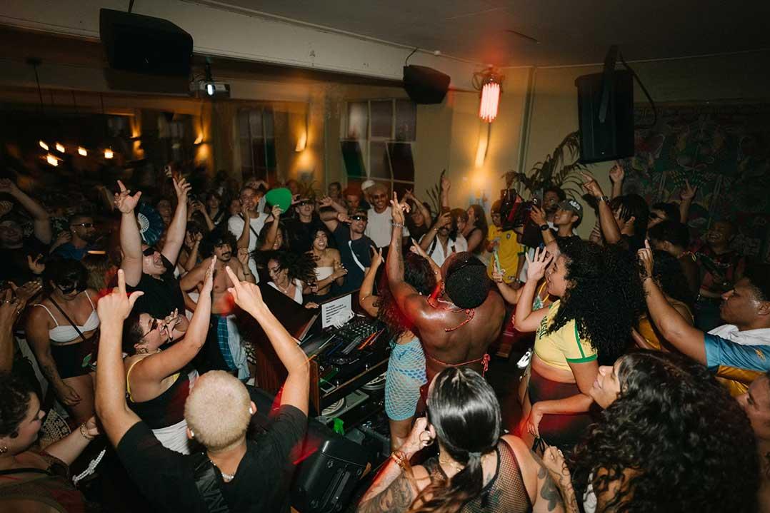 A diverse crowd dances with raised arms at a packed nightclub, surrounded by warm lighting and a DJ booth, creating an energetic, celebratory mood.