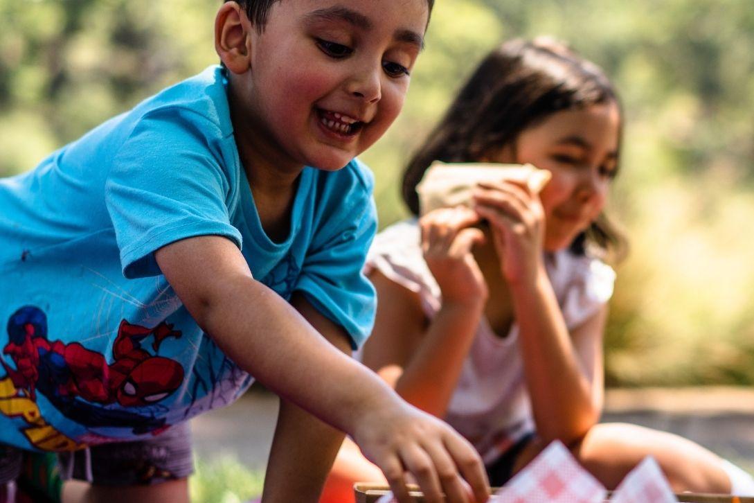 Smiling boy in a blue Spider-Man shirt reaches toward a picnic basket outdoors, while a girl in pink eats a sandwich behind him.
