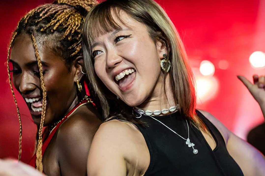 Two women dance and laugh under bright red stage lights; one wears a black top and shell choker, while the other has long braids. The mood is joyful and energetic.