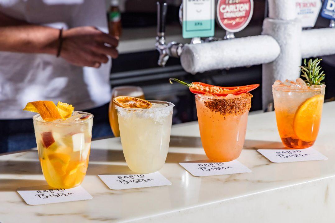 Four colorful cocktails—citrus, herb, and chili-garnished—line a bar counter beneath beer taps as a bartender works in the background, creating a lively mood.