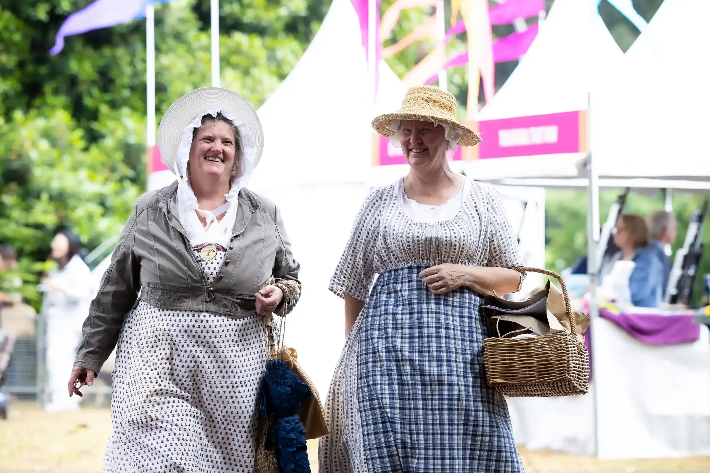 Two people dressed in historical period costumes walk along a path at an outdoor heritage festival.