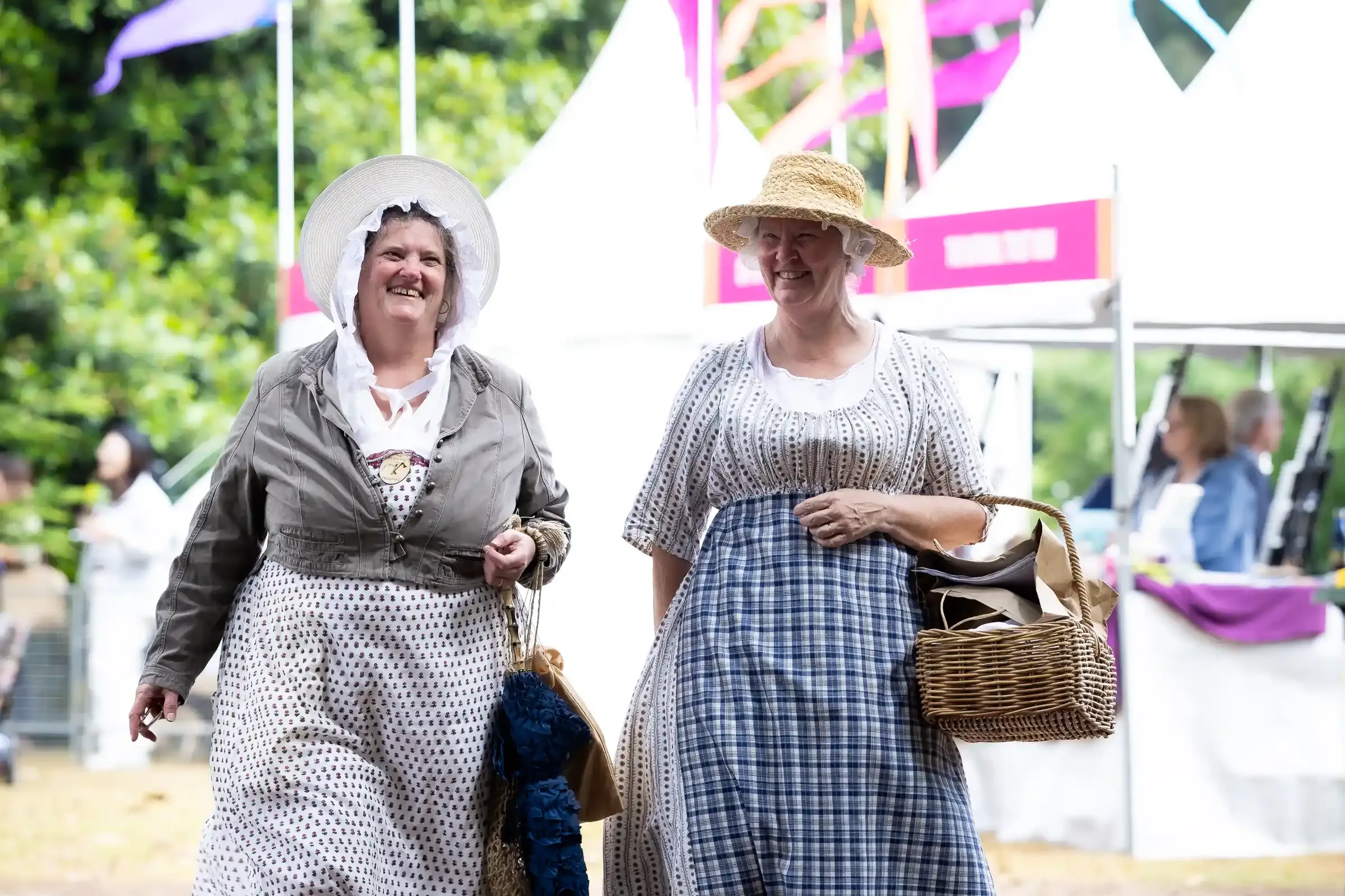 Two people dressed in historical period costumes walk along a path at an outdoor heritage festival.