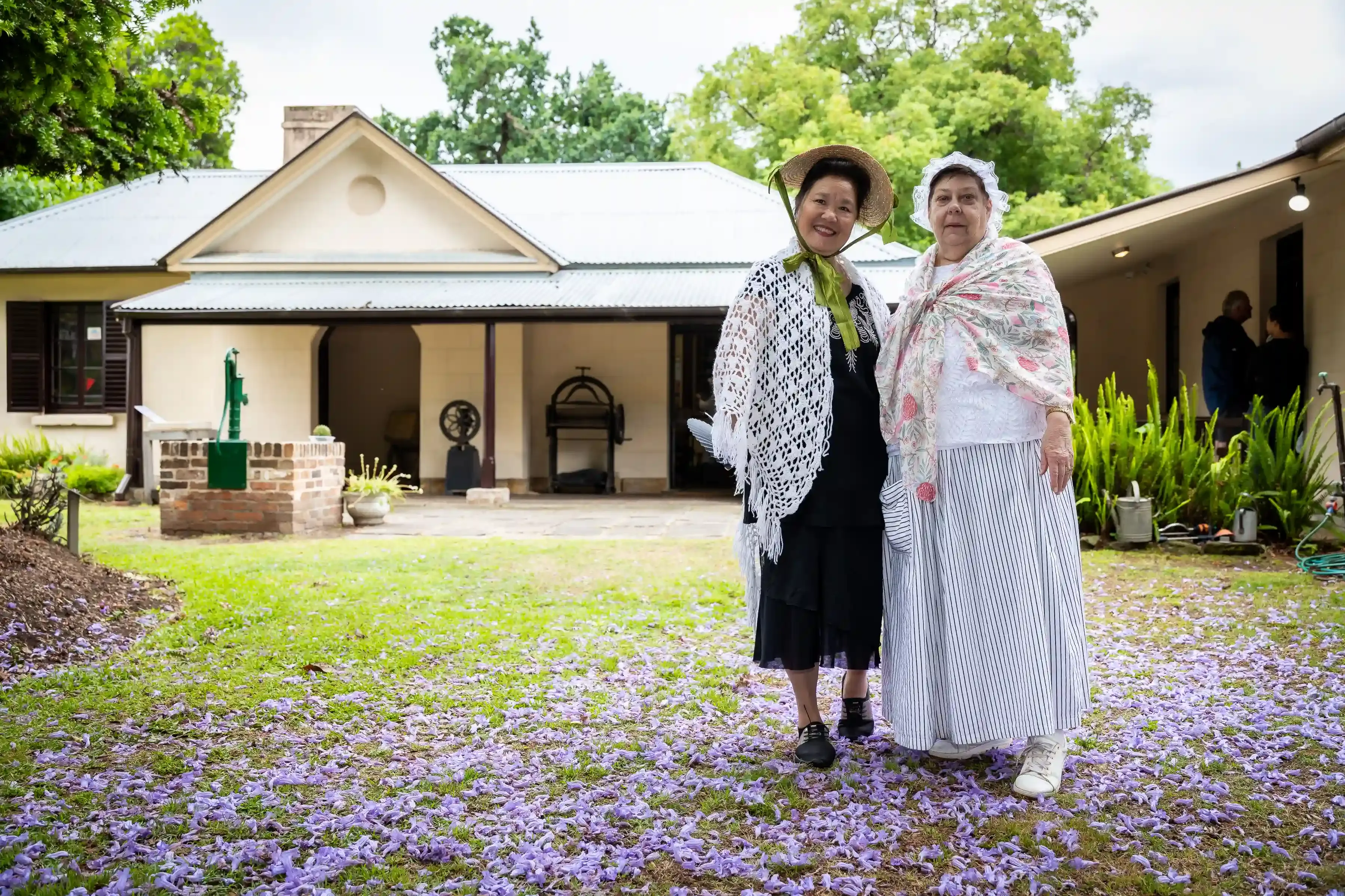 Two people dressed in historical period clothing stand in front of a heritage cottage surrounded by purple jacaranda petals.