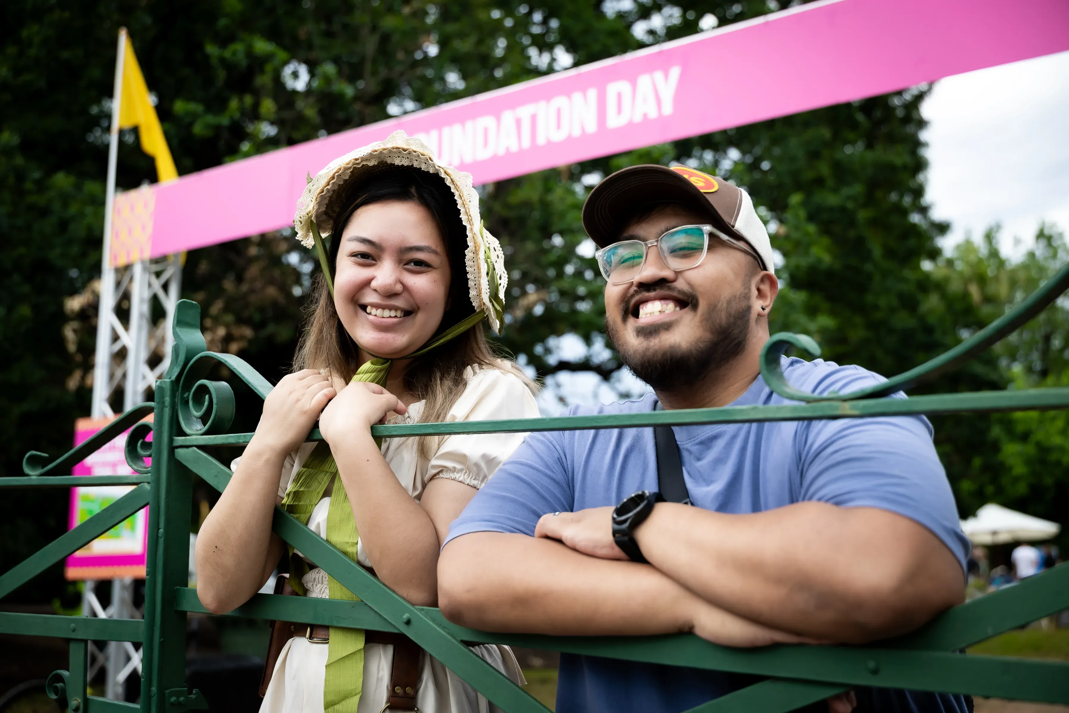 Two people lean on a green wrought-iron fence at an outdoor event, posing in front of a large pink archway with the words "Foundation Day" visible in the background.