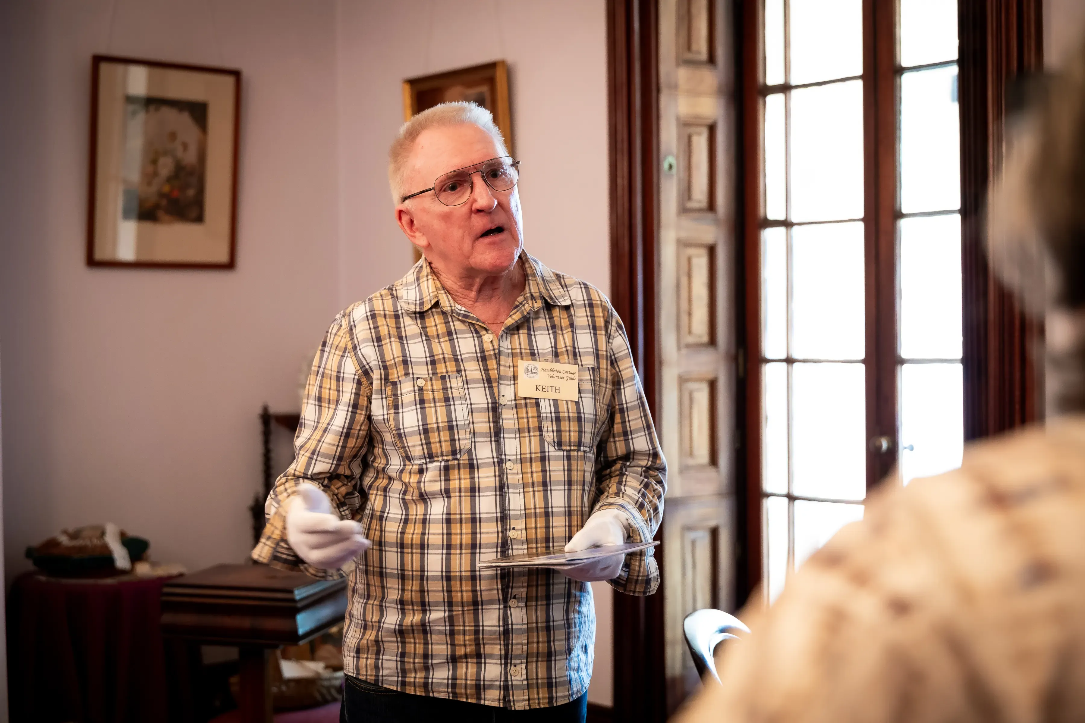A volunteer guide speaks to visitors inside a heritage house, holding a pamphlet while standing beside a tall window.