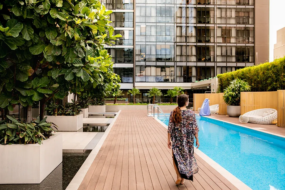 A woman walks along a wooden deck beside a bright blue rooftop pool, surrounded by lush planters and modern high-rise buildings, creating a calm urban retreat.