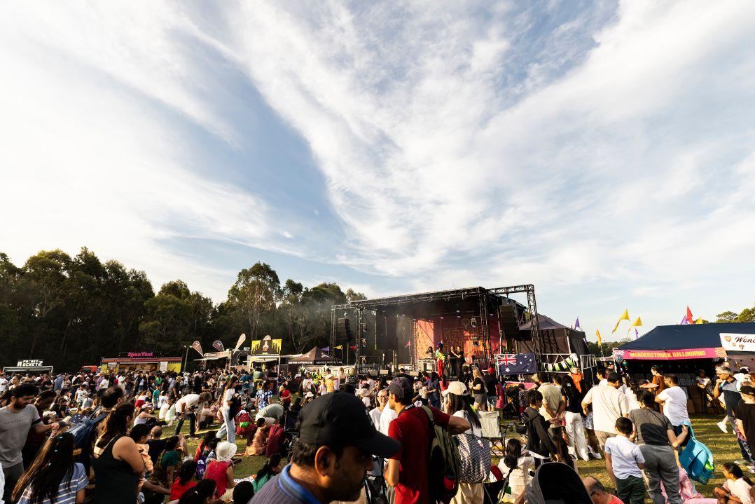 A lively outdoor music festival crowds around a stage beneath a vast, cloud-streaked sky, with colorful flags, trees, and food stalls adding to the festive atmosphere.