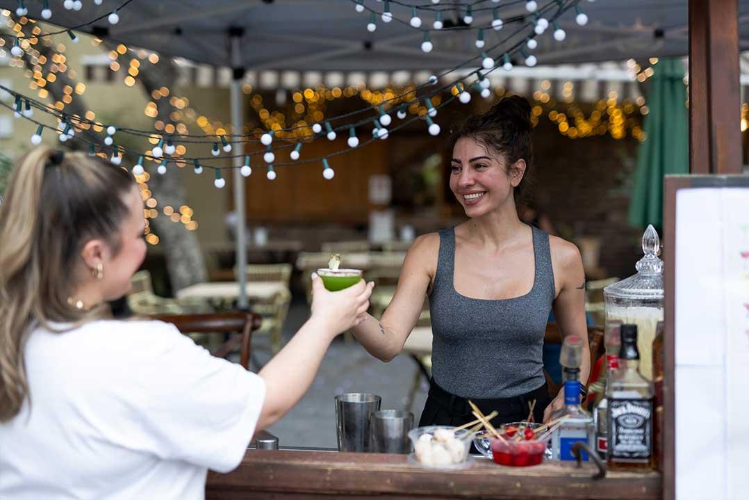A smiling bartender hands a customer a green drink at an outdoor bar decorated with string lights, creating a warm, lively evening atmosphere.