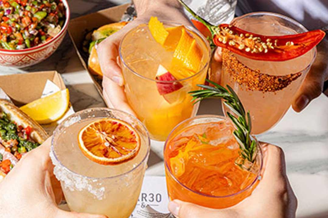 Hands raise four colorful cocktails garnished with citrus, chili, and rosemary over salsa and lime, creating a festive, summery mood.