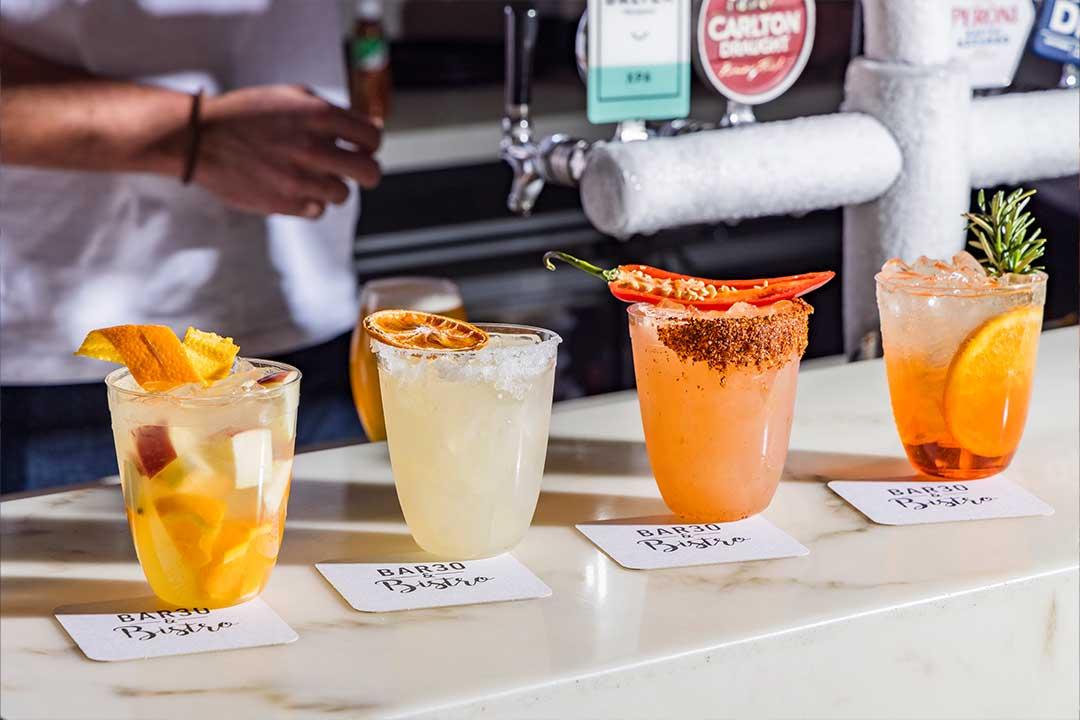 Four colorful cocktails sit on a bar counter: fruit-filled, citrus, chili-rimmed with a pepper garnish, and herb-topped, while a bartender pours in the background.