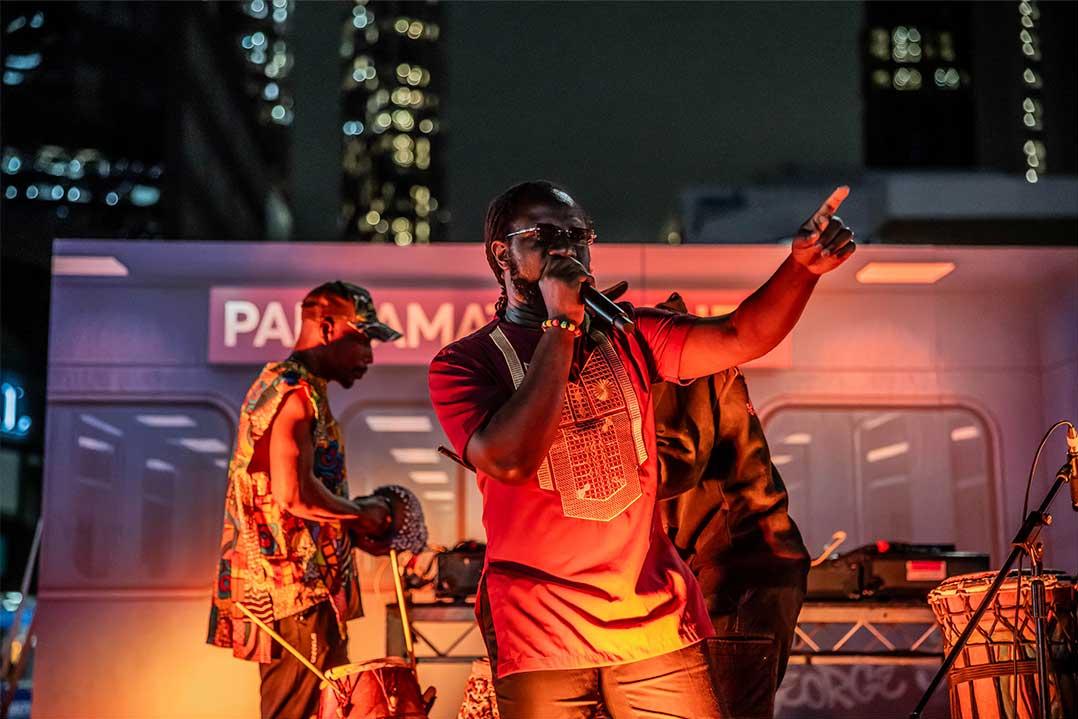 A singer in a red shirt performs into a microphone, pointing toward the crowd as musicians play behind him on a brightly lit outdoor stage at night.