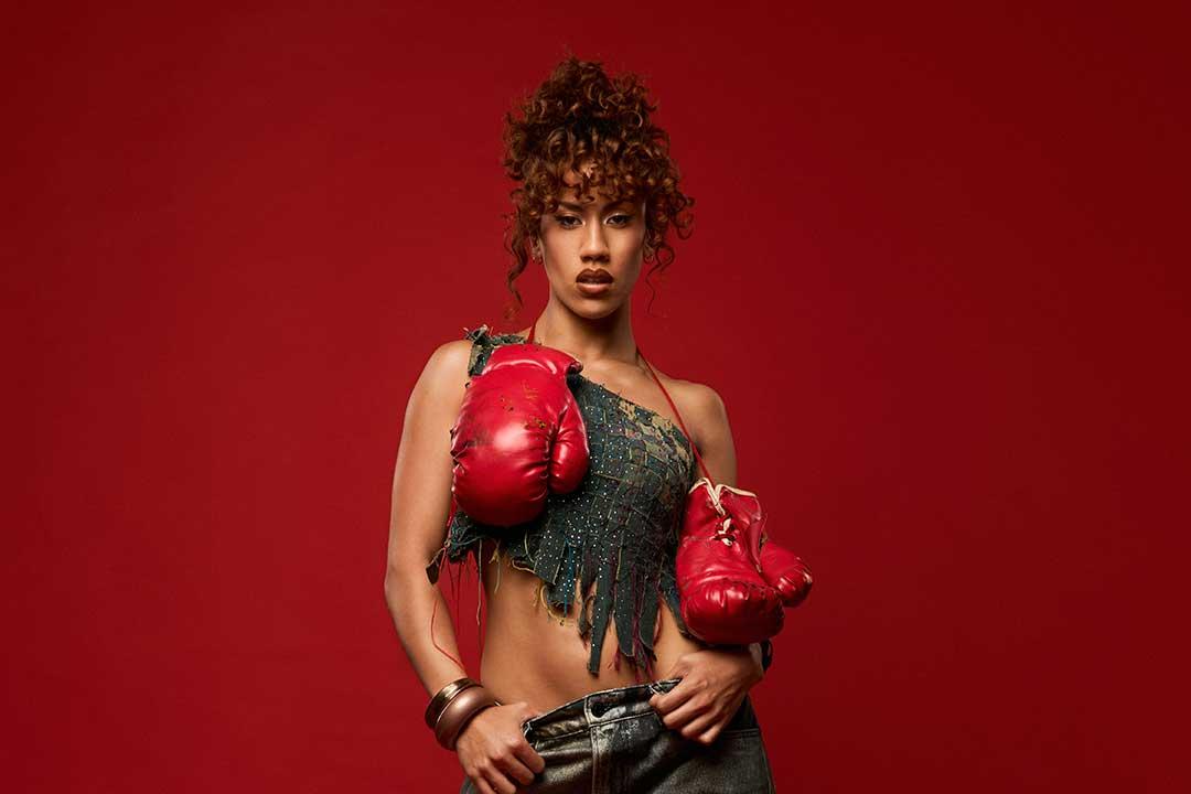 A confident model poses against a vivid red backdrop, wearing oversized red boxing gloves, a fringed dark top, and curly hair in an updo.