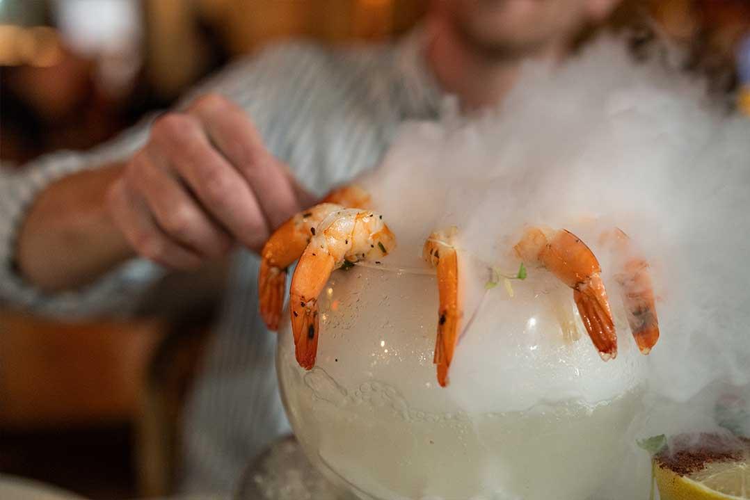 A chef arranges orange shrimp atop a mound of ice as vapor rises, creating an elegant seafood display in a warmly lit restaurant.
