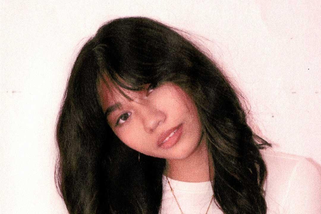 A close-up portrait of a woman with long dark hair tilting her head and smiling softly, wearing a light top against a pale pink backdrop; warm, dreamy tone.