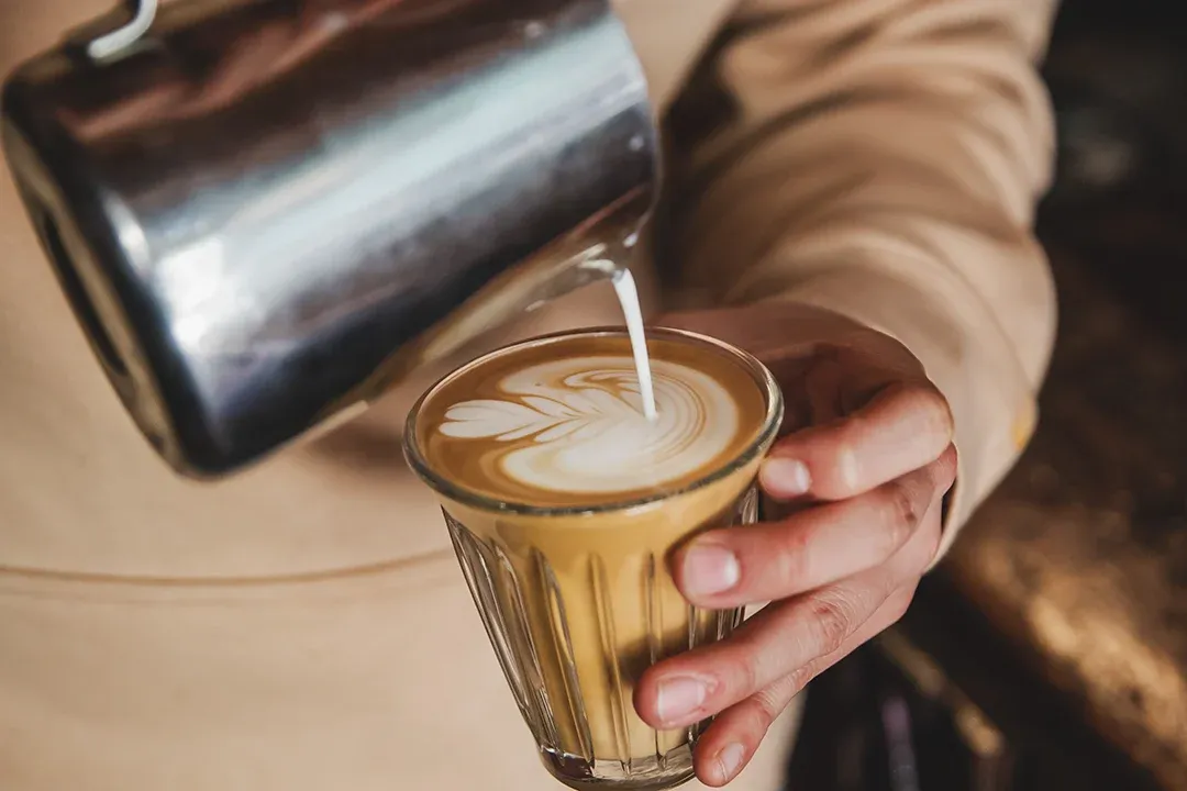 A barista pours steamed milk from a metal pitcher into a glass of espresso, creating latte art; the warm close-up feels calm and inviting.