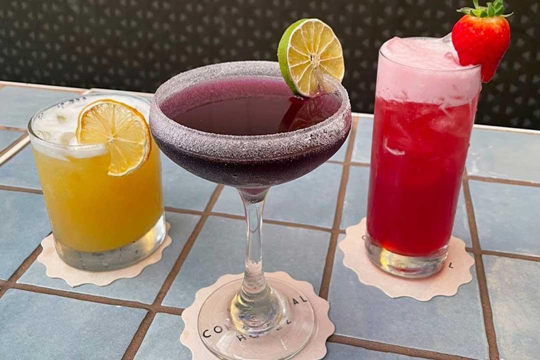 Three colorful cocktails—a yellow citrus drink, dark berry wine, and pink strawberry drink—sit on a tiled table, garnished with lemon and strawberry, creating a festive mood.