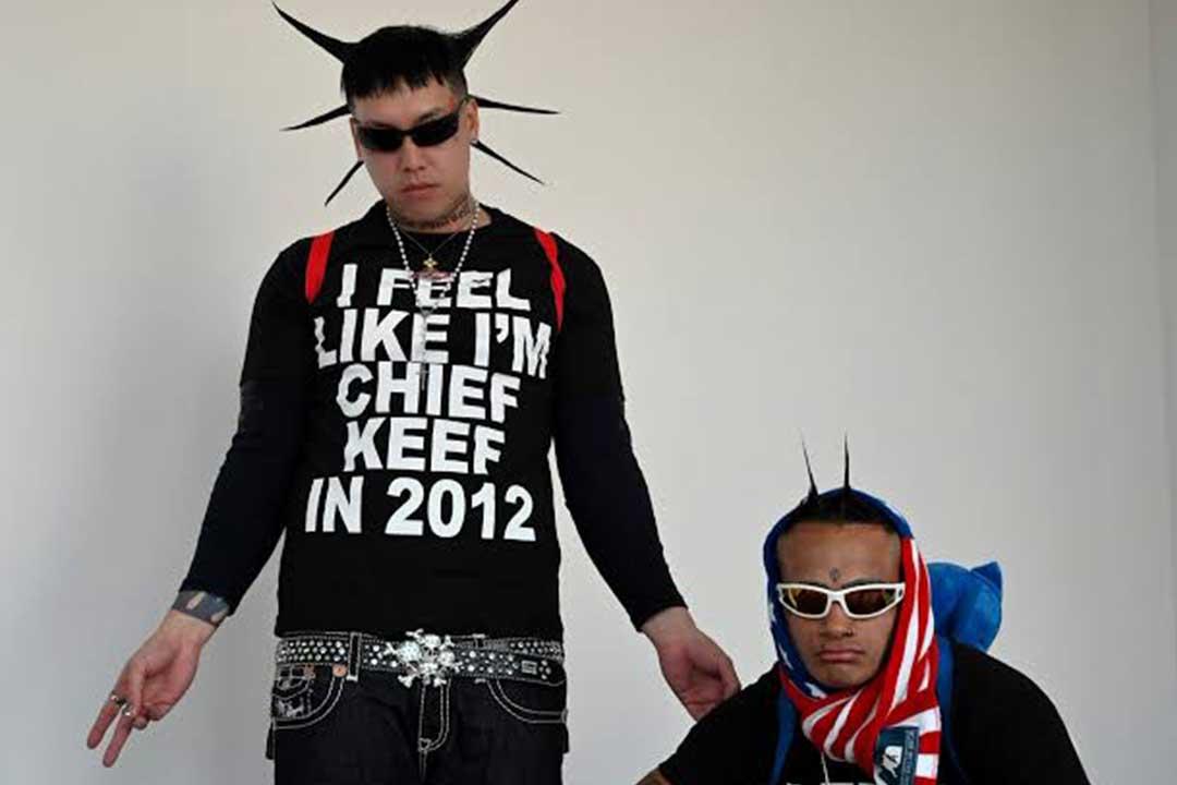 Two men in a studio pose in bold streetwear: one wears spiked hair and a “Feel Like I’m Chief Keef in 2012” shirt; the other sports shades and a flag scarf.