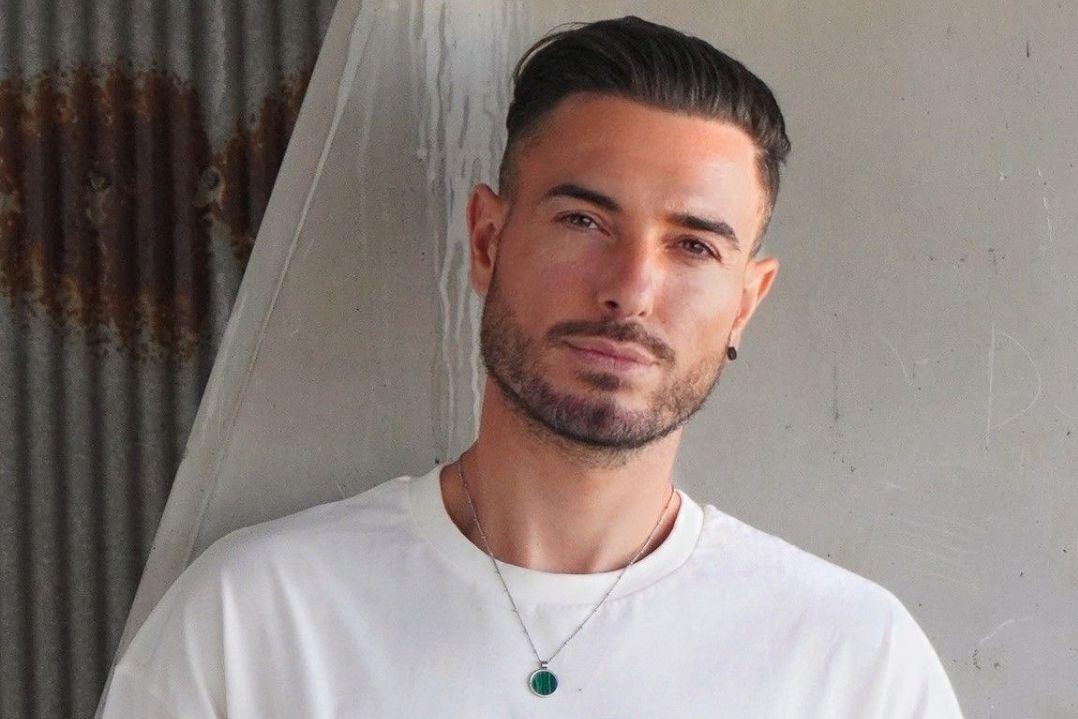 Portrait of a young man with short dark hair and a trimmed beard, wearing a white shirt and pendant necklace against a weathered wall; calm, confident expression.