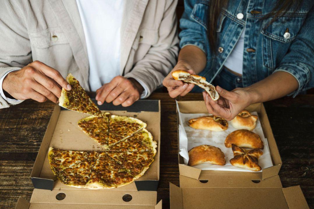 Two people share a meal: one lifts a slice of herb-topped pizza from a box while the other holds a piece of bread beside a box of pastries.