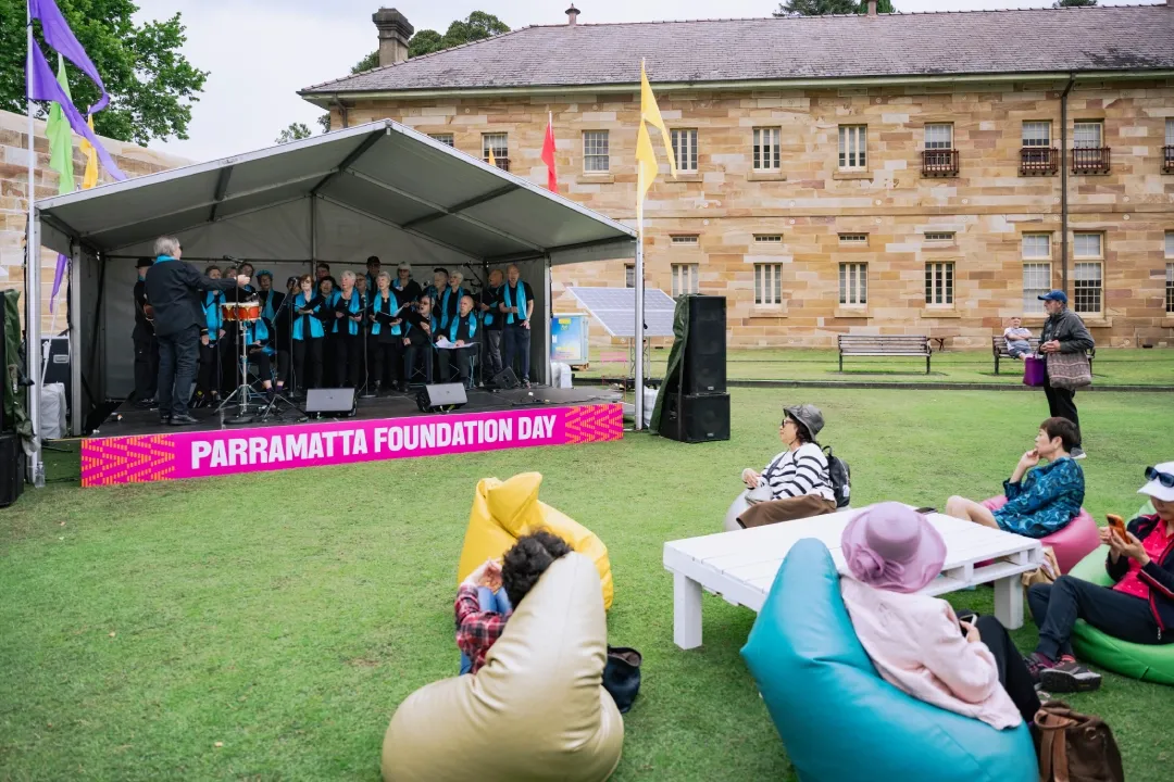 A choir performs on an outdoor stage during Parramatta Foundation Day, with audience members seated on bean bags and picnic furniture on the lawn.