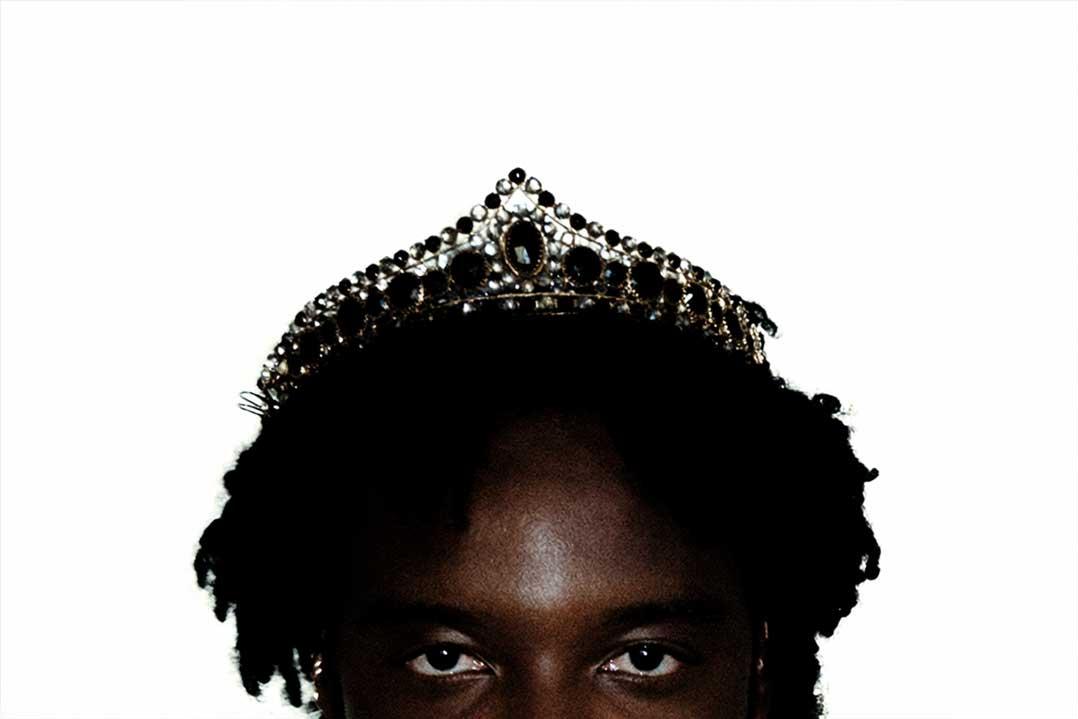 Close-up of a dark-skinned person with natural curls wearing an ornate jeweled tiara, eyes visible against a plain white background; regal and striking.
