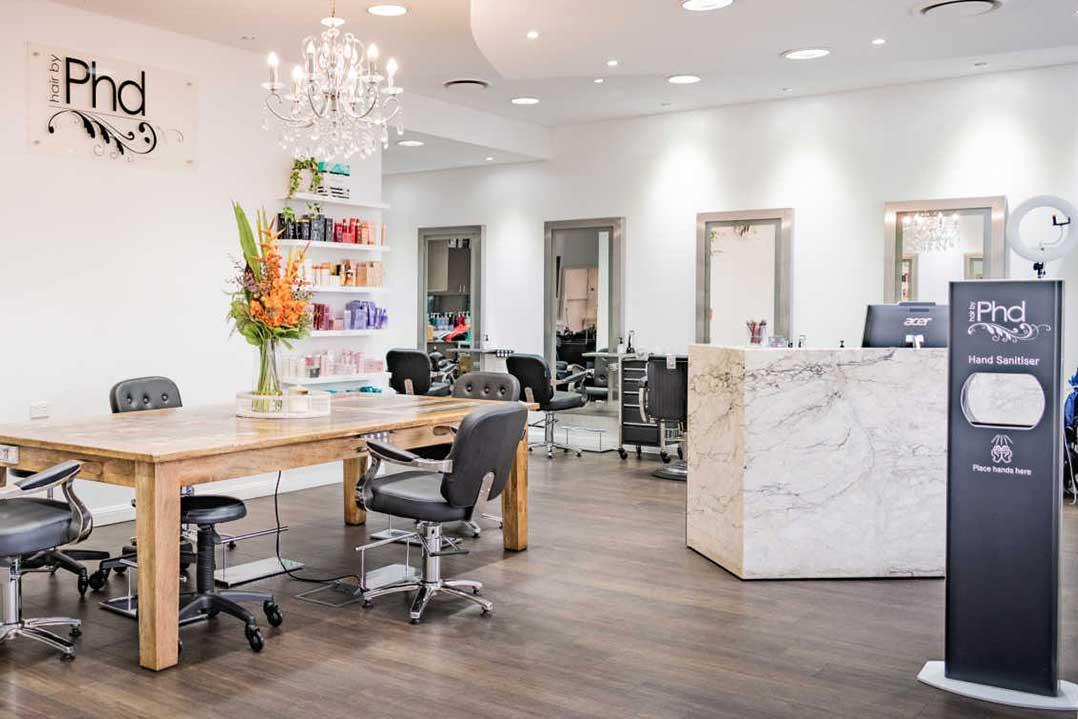 Bright, modern PhD nail salon with manicure tables, colorful polish shelves, flowers, a marble reception desk, and sanitizer stand; clean, calm atmosphere.
