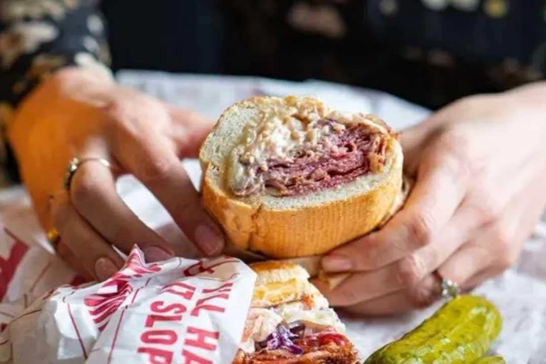 Hands hold a sliced crusty sandwich filled with cured meat, beside a wrapped sandwich and pickle; the close-up feels warm and appetizing.