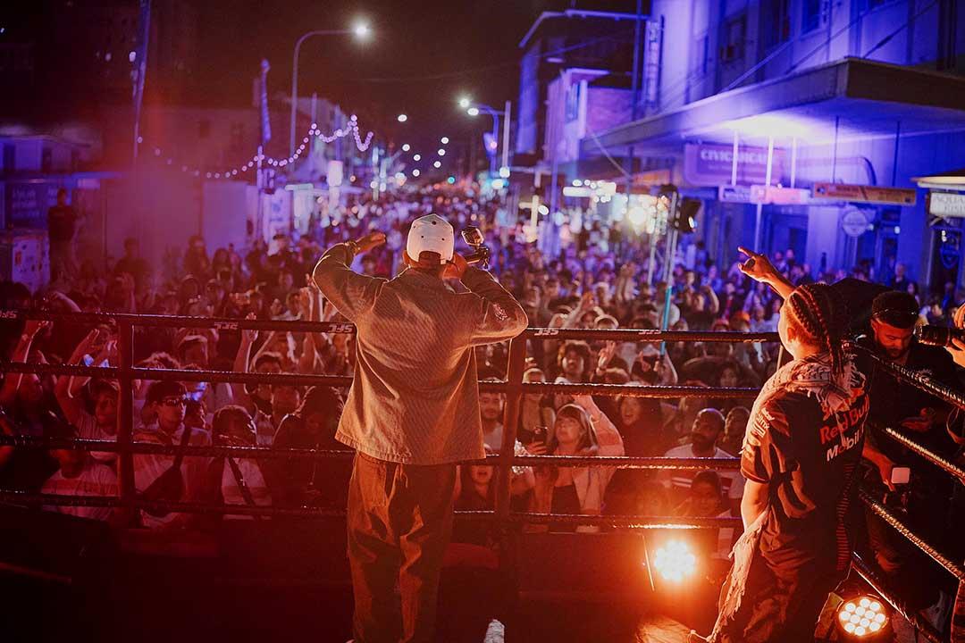 A cap-wearing performer raises his arms toward a packed nighttime street crowd as a dancer points beside him under vivid red and blue stage lights.