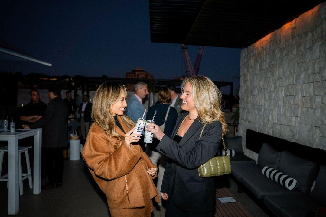 Two people stand outdoors at an evening social gathering, raising canned drinks in a toast.