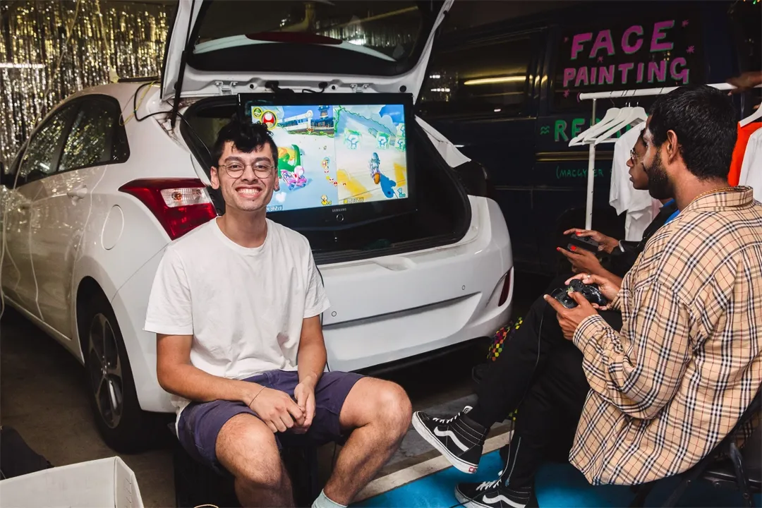 At a lively night event, a smiling young man sits beside an open SUV trunk displaying a colorful game, while another man chats nearby under bright lights.