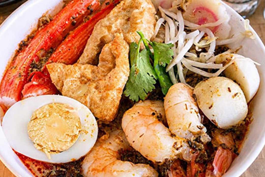 A vibrant bowl of spicy seafood noodle soup topped with crab legs, shrimp, squid, boiled egg, bean sprouts, fish cake, and fresh cilantro.