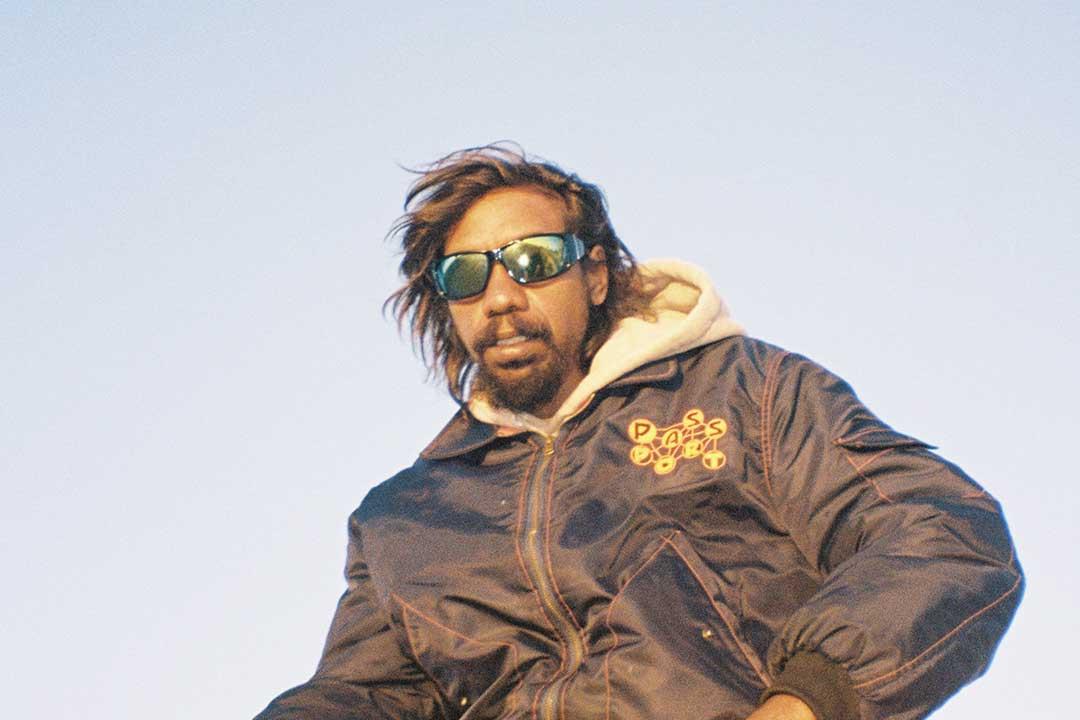 A man in reflective sunglasses and a dark jacket over a cream hoodie looks down against a pale sky, conveying a relaxed, confident streetwear style.