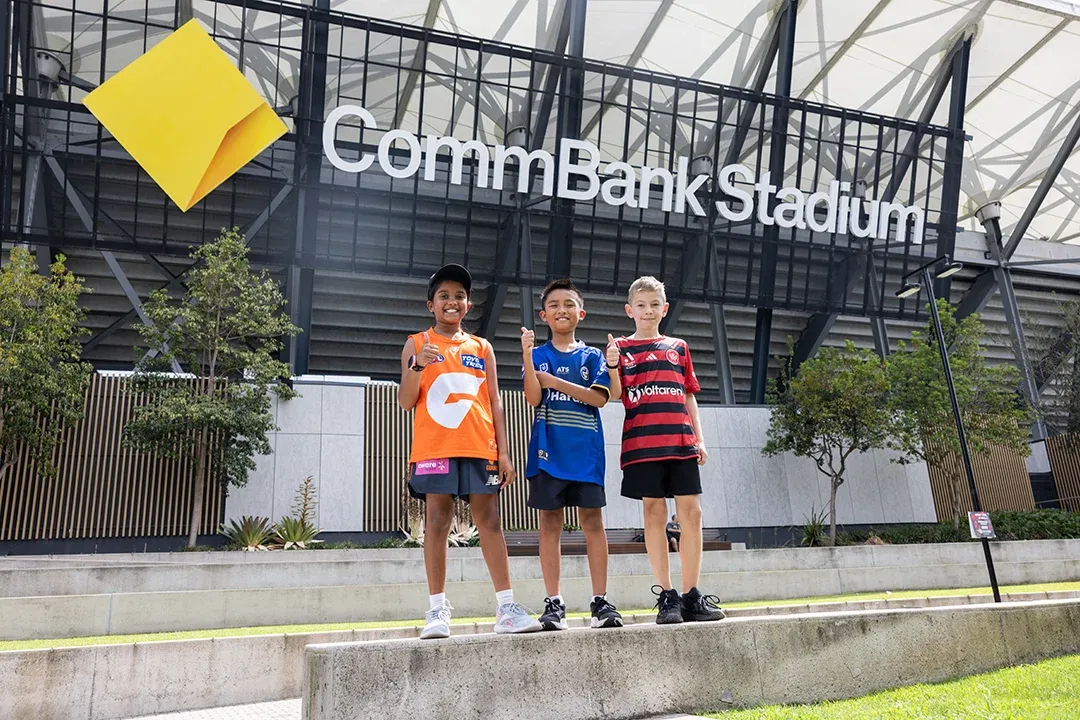 Three young soccer fans in orange, blue, and red-and-black jerseys pose outside CommBank Stadium beneath its sign, smiling proudly on a bright day.