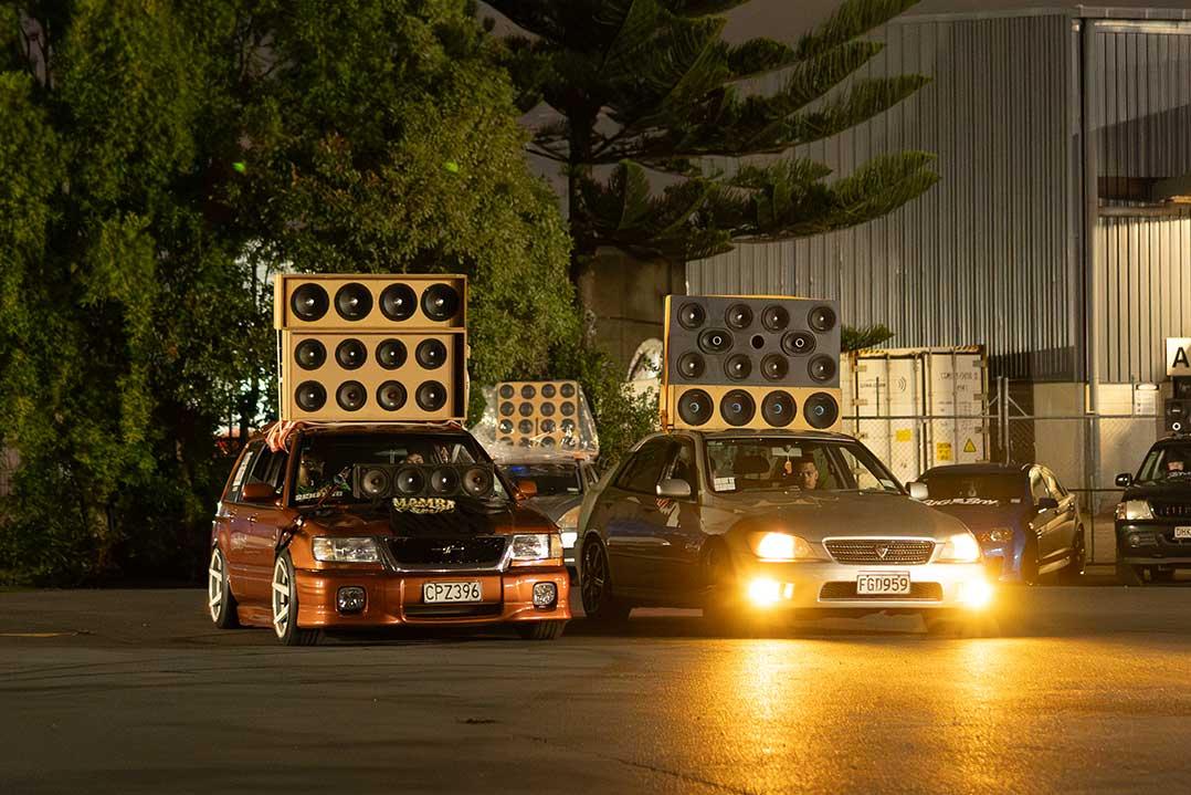 Two modified cars with towering speaker stacks sit at night outside an industrial building; warm headlights and streetlights create a dramatic car-audio showcase.