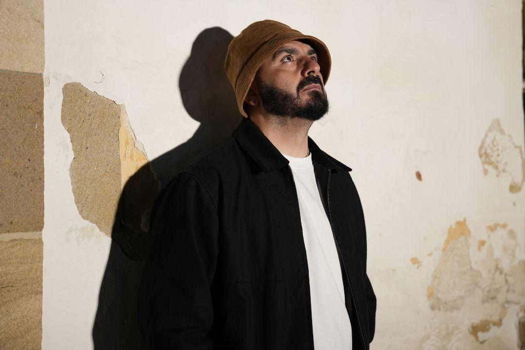 A bearded man in a brown bucket hat and black coat looks upward against a worn, peeling wall, casting a sharp shadow; contemplative, moody portrait.