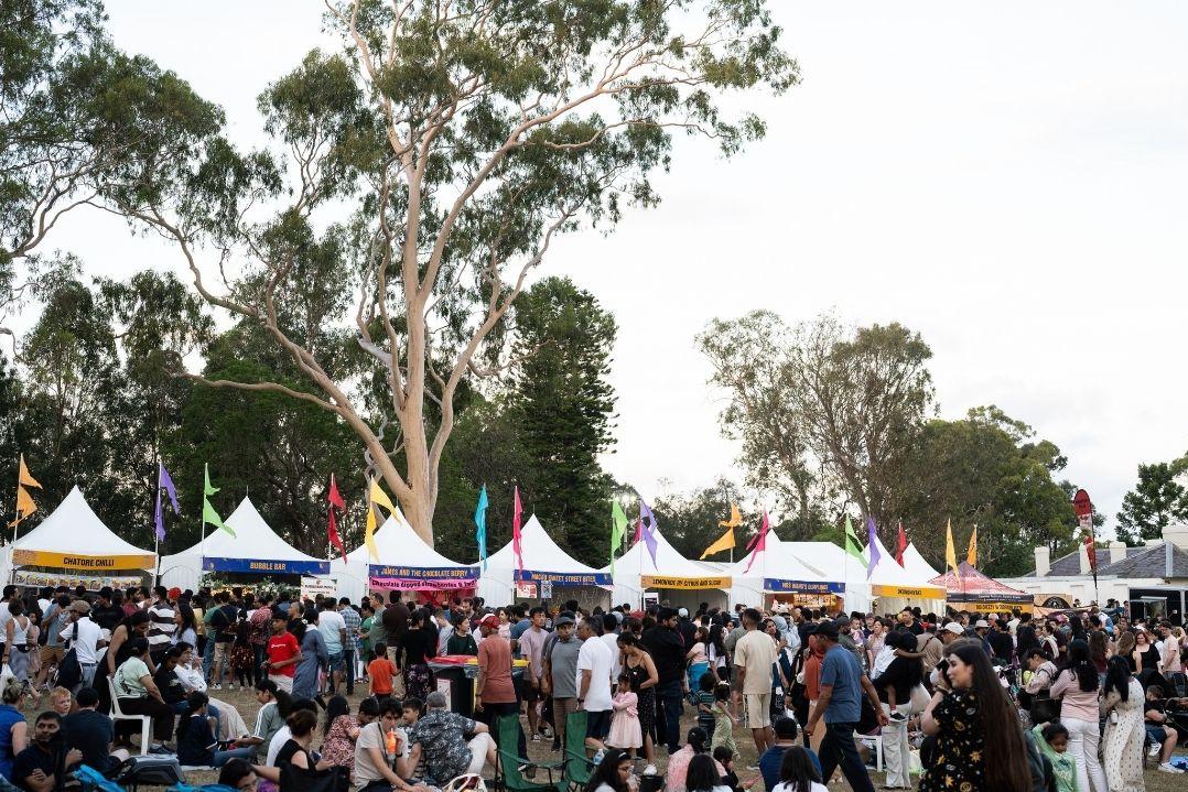 A busy outdoor festival fills a tree-lined park, with crowds browsing white market tents and colorful flags overhead, creating a lively, relaxed mood.