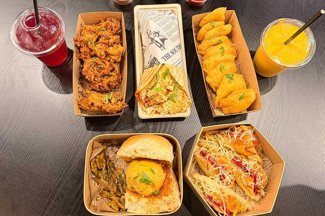 Overhead view of colorful Indian street food in takeaway trays—fried snacks, sandwiches, noodles, and two bright drinks—looks lively and inviting.