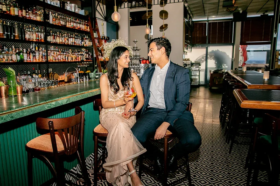 A couple in formal attire sit close together at a stylish, bottle-lined bar, smiling at each other over drinks in warm, intimate light.