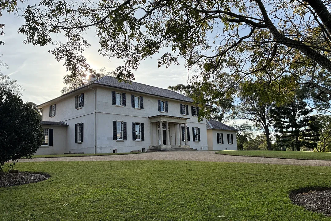 A stately white two-story colonial home with black shutters stands on a broad green lawn beneath spreading trees, creating a calm, historic feel.